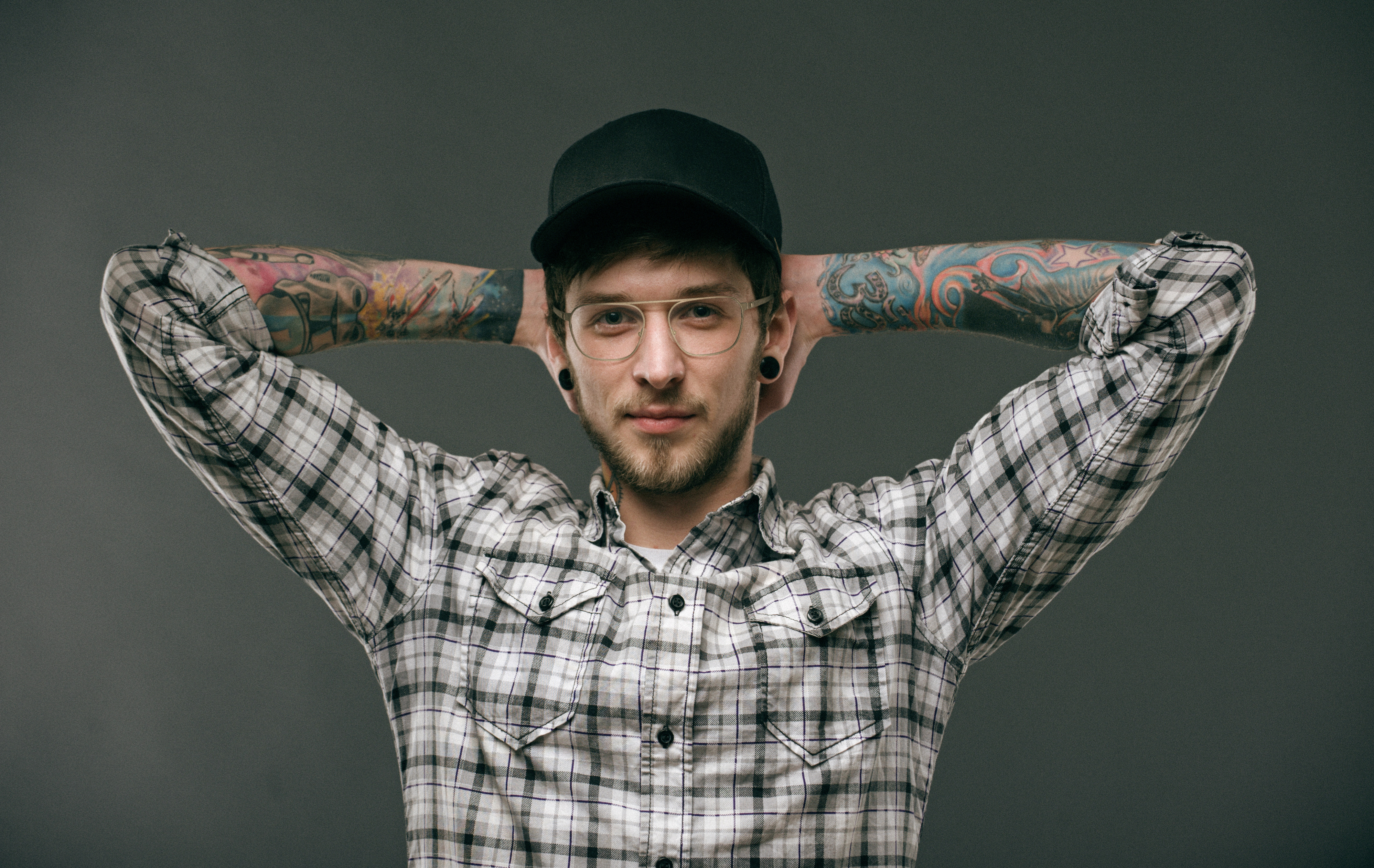 Homme avec tatouage sur l'avant-bras, casquette et lunettes métal fines,mains derrière la tête.